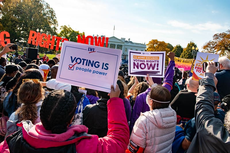 A crowd gathers outdoors holding signs advocating for voting rights, including one that reads "Voting is People Power," near the White House.
