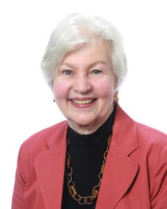 Elderly woman with short white hair, wearing a coral blazer, black turtleneck, and beaded necklace, smiling in front of a white background.