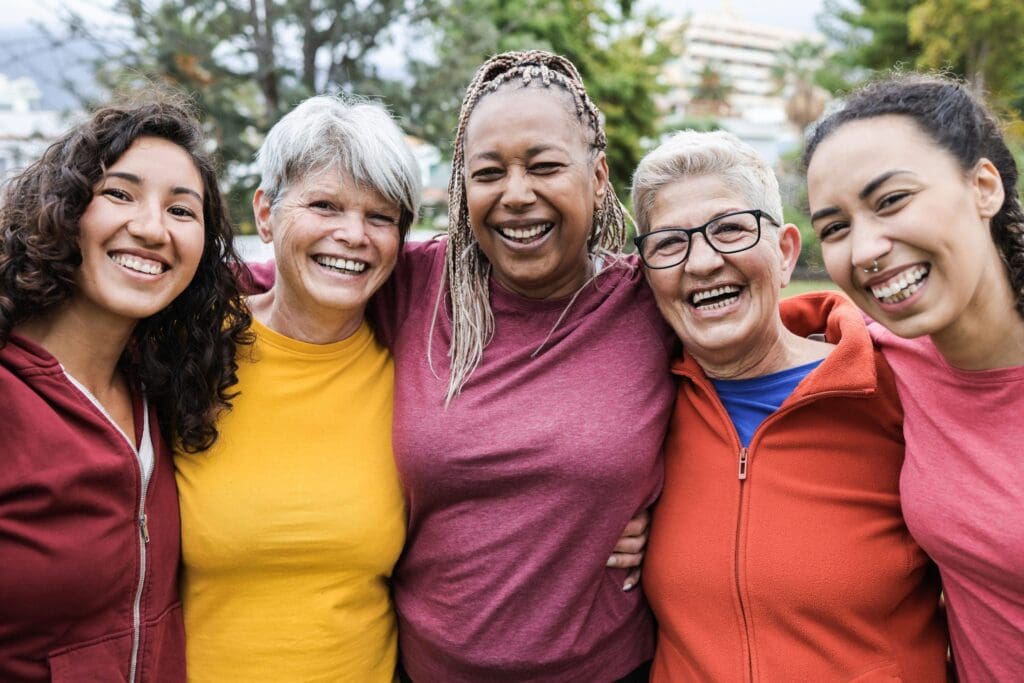 Five women of different ages stand close together outdoors, smiling at the camera, dressed in casual colorful clothing.
