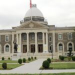 A large, multi-story stone building with a central dome and columns, featuring a statue of a person on the front lawn.