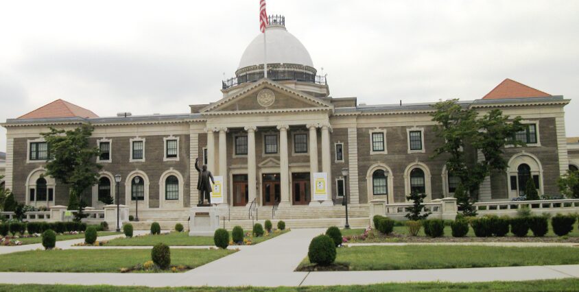 A large, multi-story stone building with a central dome and columns, featuring a statue of a person on the front lawn.