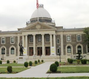 A large, multi-story stone building with a central dome and columns, featuring a statue of a person on the front lawn.